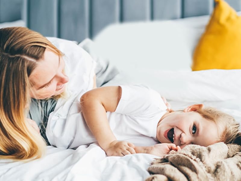 Adulto e bambino su letto bianco. Cuscino giallo con logo verde visibile. Scena diurna in camera da letto raffigurante comfort e riposo.